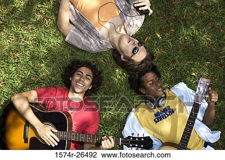 High angle view of two young men lying on grass and playing guitars with a young man listening to music View Large Photo Image Stock Image - High angle view of two young men lying on grass and playing guitars with a young man listening to music. Fotosearch
