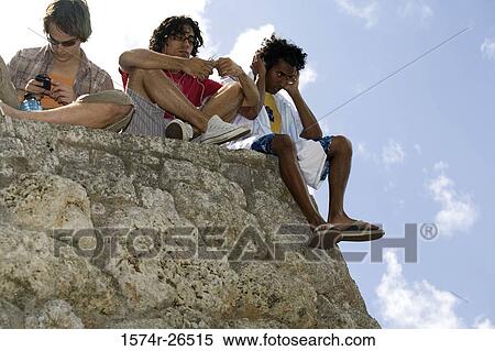 Stock Photography - Low angle view of three young men sitting on a rock. Fotosearch