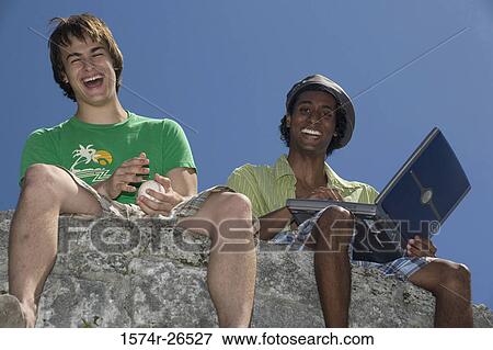 Low angle view of two young men sitting on a rock View Large Photo Image Stock Photo - Low angle view of two young men sitting on a rock. Fotosearch