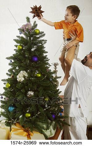 Stock Photo - Mature man lifting his son to decorate a Christmas tree. Fotosearch