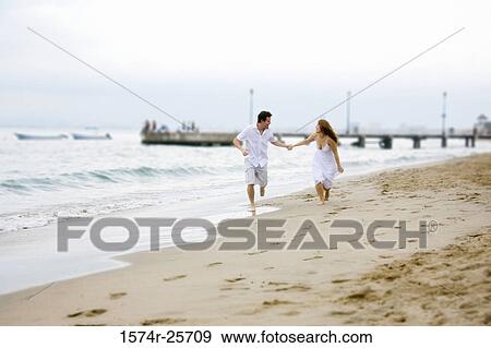 Stock Photo - Mid adult man and a young woman holding each other's hands and running on the beach. Fotosearch
