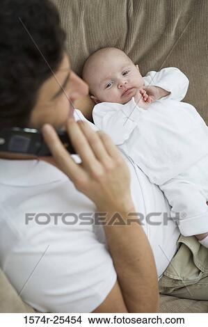 Mid adult man holding his baby while talking on a mobile phone View Large Photo Image Picture - Mid adult man holding his baby while talking on a mobile phone. Fotosearch