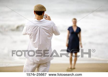 Rear view of a mid adult man taking a picture of a young woman with a digital camera on the beach View Large Photo Image Stock Photo - Rear view of a mid adult man taking a picture of a young woman with a digital camera on the beach. Fotosearch