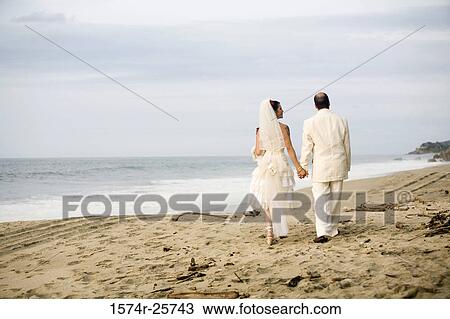 Rear view of a newlywed couple holding hands and walking on the beach View Large Photo Image Stock Image - Rear view of a newlywed couple holding hands and walking on the beach. Fotosearch