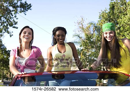 Stock Image - Two teenage girls and a young woman standing in a convertible car. Fotosearch