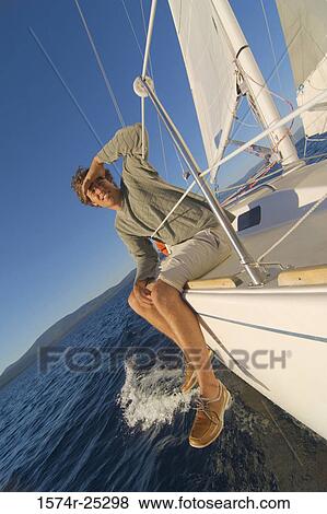 Stock Photo - Young man sitting on the edge of a sailboat and shielding his eyes. Fotosearch