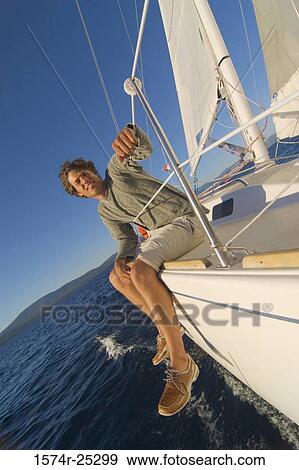 Stock Photo - Young man sitting on the edge of a sailboat and smiling. Fotosearch