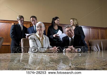 Stock Image - Businessman and businesswoman sitting by conference table while colleagues discussing in background. Fotosearch