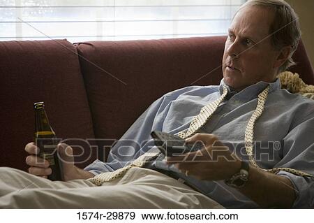 Businessman lying on sofa holding a remote control and a beer bottle View Large Photo Image Stock Photo - Businessman lying on sofa holding a remote control and a beer bottle. Fotosearch
