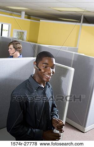 Businessman using headset in an office with colleague in the background View Large Photo Image Stock Photo - Businessman using headset in an office with colleague in the background. Fotosearch