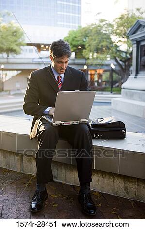 Stock Image - Businessman using laptop computer by sidewalk. Fotosearch