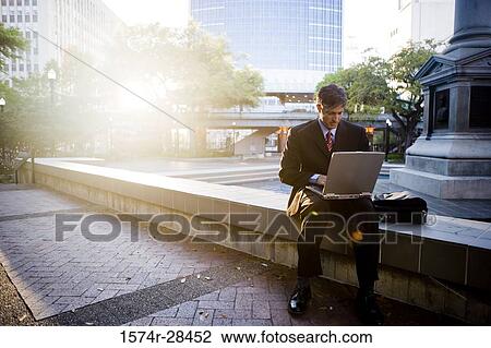 Stock Image - Businessman using laptop computer by sidewalk. Fotosearch