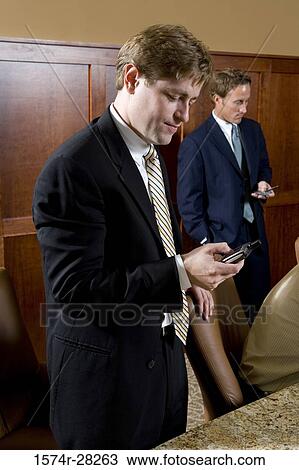 Stock Image - Businessmen text messaging on mobile phone in an office. Fotosearch