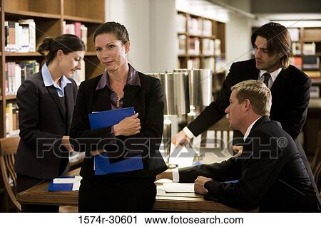 Stock Image - Businesswoman standing by table while colleagues discussing in background. Fotosearch