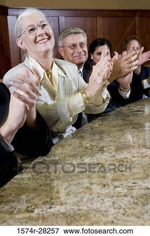 Stock Photo - Cheerful business people sitting by conference table and applauding in an office. Fotosearch