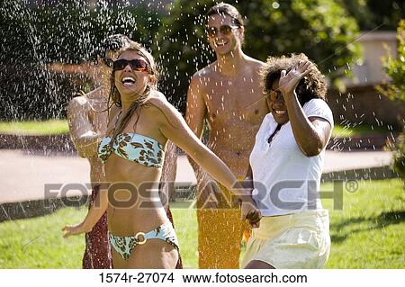 Cheerful friends enjoying with water sprinkler on lawn View Large Photo Image Picture - Cheerful friends enjoying with water sprinkler on lawn. Fotosearch