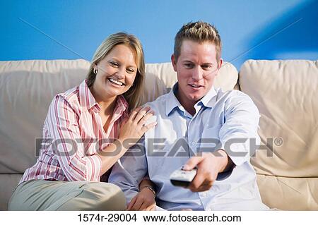 Cheerful mid adult couple with remote control sitting on a sofa View Large Photo Image Picture - Cheerful mid adult couple with remote control sitting on a sofa. Fotosearch