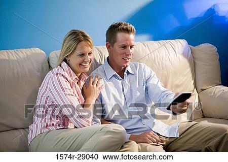 Stock Image - Cheerful mid adult couple with remote control sitting on a sofa. Fotosearch