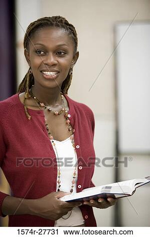Stock Image - Cheerful teacher holding book and standing in the classroom. Fotosearch