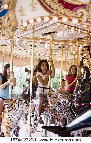 Stock Photo - Cheerful teenage girls sitting on a carousel and waving hands. Fotosearch