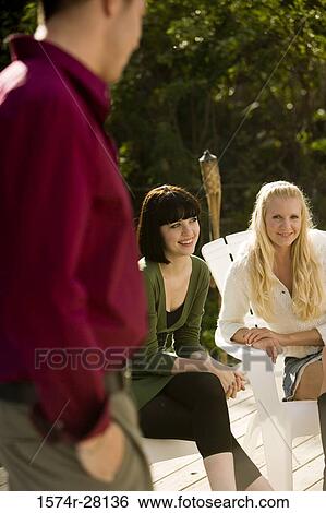 Cheerful women sitting on armchairs while young man standing with hands in pockets View Large Photo Image Stock Photograph - Cheerful women sitting on armchairs while young man standing with hands in pockets. Fotosearch