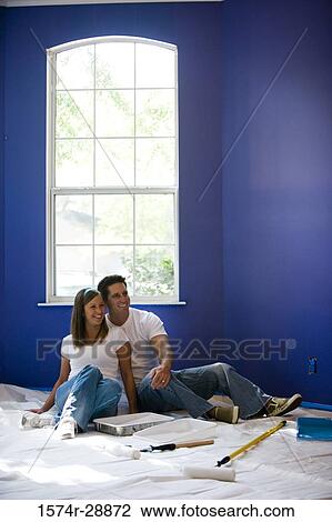 Stock Image - Cheerful young couple sitting by paint tray and paint roller in house. Fotosearch