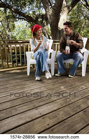 Stock Photograph - Cheerful young couple sitting together on armchairs and eating. Fotosearch