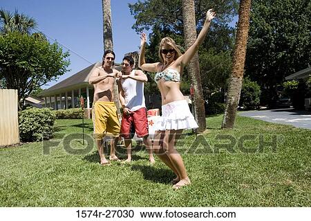 Stock Image - Cheerful young woman dancing on lawn while young men standing in the background. Fotosearch
