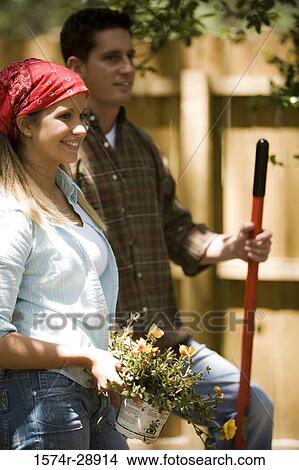 Cheerful young woman holding plant while young man standing with shovel View Large Photo Image Picture - Cheerful young woman holding plant while young man standing with shovel. Fotosearch