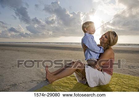 Cheerful young woman sitting with her son at the beach View Large Photo Image Stock Image - Cheerful young woman sitting with her son at the beach. Fotosearch