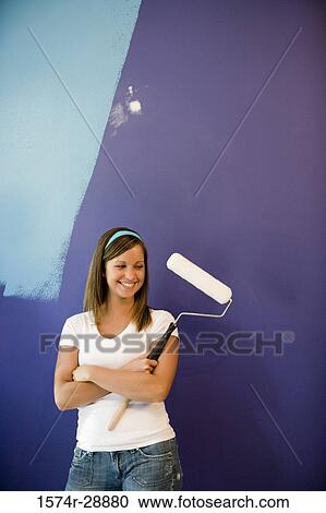 Stock Image - Cheerful young woman with paint roller by wall. Fotosearch