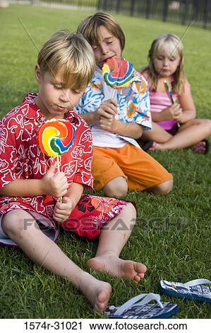 Stock Image - Children sitting on grass and eating lollipop. Fotosearch