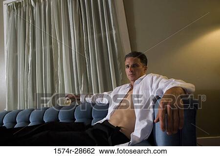 Stock Image - Contemplative mid adult man holding a glass while sitting on sofa. Fotosearch