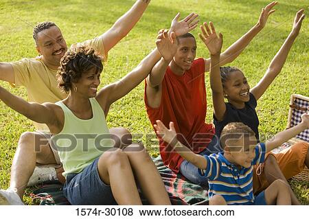 Stock Photo - Family sitting with arms raised on blanket at park. Fotosearch
