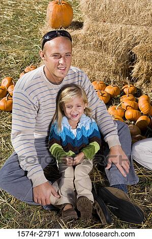 Stock Image - Father sitting with his daughter on grass by straw bundle. Fotosearch