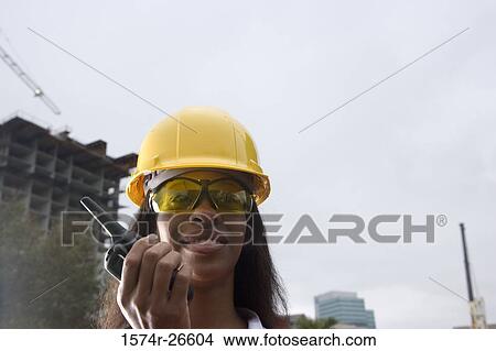 Picture - Female architect wearing hard hat and holding walkie-talkie against sky. Fotosearch