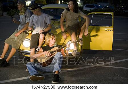 Friends using mobile phones and playing guitar by a car at the parking area View Large Photo Image Stock Image - Friends using mobile phones and playing guitar by a car at the parking area. Fotosearch
