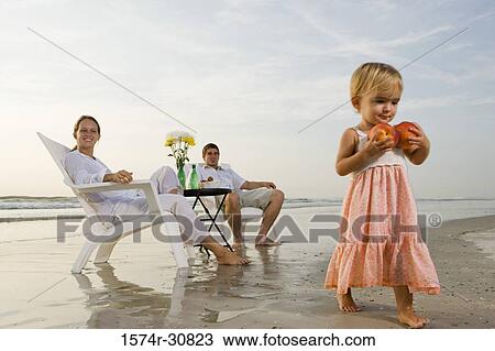Girl carrying apples with parents sitting in the background on the beach View Large Photo Image Stock Image - Girl carrying apples with parents sitting in the background on the beach. Fotosearch