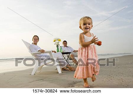 Girl carrying fruit with parents sitting in the background on the beach View Large Photo Image Picture - Girl carrying fruit with parents sitting in the background on the beach. Fotosearch