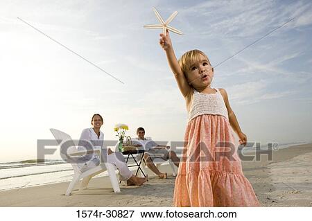 Girl holding starfish with parents sitting in the background on the beach View Large Photo Image Stock Photo - Girl holding starfish with parents sitting in the background on the beach. Fotosearch