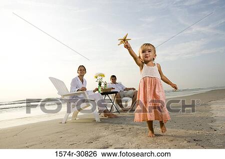 Girl holding starfish with parents sitting in the background on the beach View Large Photo Image Stock Photograph - Girl holding starfish with parents sitting in the background on the beach. Fotosearch