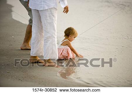 Stock Image - Girl playing in water with parents standing beside at beach. Fotosearch