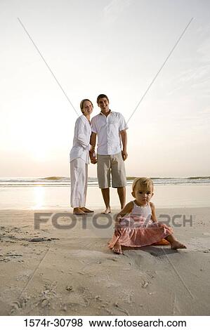 Stock Photo - Girl sitting on the beach with parents in the background. Fotosearch