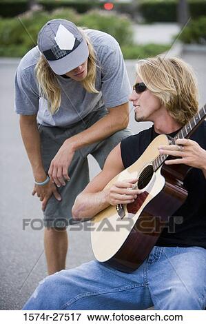 Stock Photo - Man playing a guitar while conversing with his friend. Fotosearch