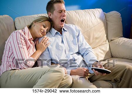 Stock Image - Mid adult couple with remote control relaxing on sofa. Fotosearch
