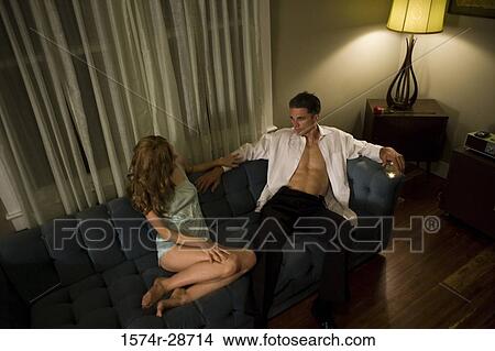 Picture - Mid adult man sitting with a young prostitute on sofa. Fotosearch