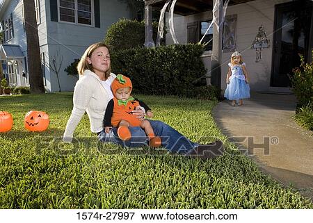 Stock Photo - Mother sitting with her son in lawn while daughter in costume standing in the background. Fotosearch