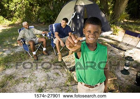 Stock Image - Portrait of a boy holding bait hook while men discussing by tent at forest. Fotosearch