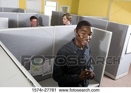 Stock Image - Portrait of a businessman leaning by desk while colleagues conversing in the background. Fotosearch