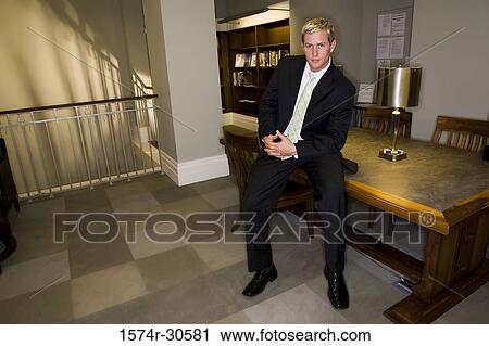 Stock Image - Portrait of a businessman sitting on table in an office. Fotosearch